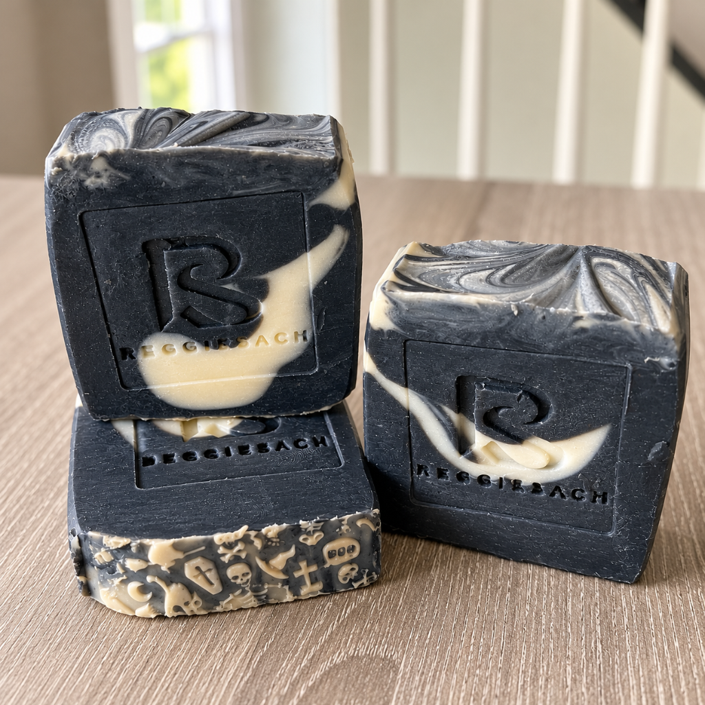ReggieSach Spooky Vanilla Bar Soap, black cold process bars with cream swirls and embossed ReggieSach logo stacked on a wood surface with a Halloween edge pattern