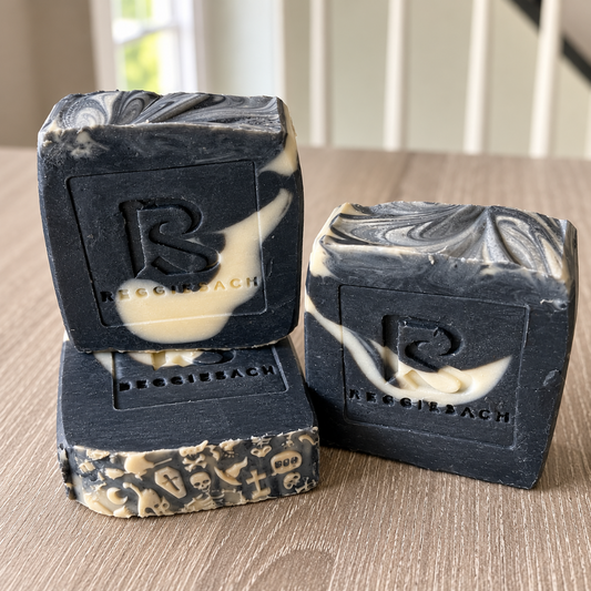 ReggieSach Spooky Vanilla Bar Soap, black cold process bars with cream swirls and embossed ReggieSach logo stacked on a wood surface with a Halloween edge pattern